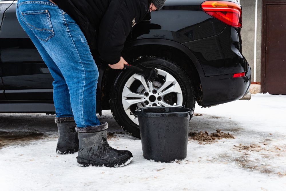 Muškarac u zimskoj odeći čisti zadnji točak crnog automobila na snegu